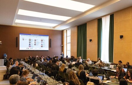 approximately 50 people in a large meeting room with connection to others online through screen on the wall